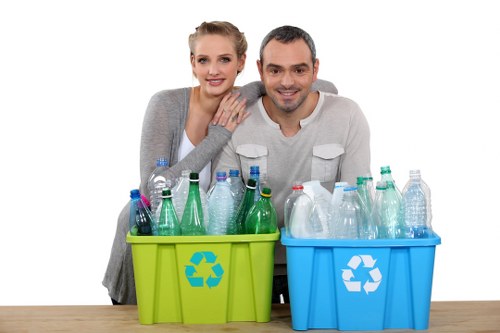 A smiling young couple with fair skin and light-colored hair, positioned behind two small recycling bins on a wooden surface, with the woman resting her arms on the man's shoulder. The bins are bright green and blue, each featuring a white recycling symbol on the front, and filled with an assortment of empty plastic bottles of various sizes and colors, including clear, green, and blue. The background is plain white, highlighting the focus on the recycling containers and the couple. The scene suggests environmentally conscious waste separation, consistent with services provided by Commercial Waste Removal Penge for domestic or commercial waste management in the local area. The plastic bottles appear clean and ready for collection or recycling, emphasizing the importance of responsible rubbish disposal and recycling practices in the community. The lighting is even and professional, with no shadows or distractions, creating a clear, neutral visual suitable for a rubbish removal website or environmental awareness context, supporting local SEO for waste management services in Penge and surrounding areas.