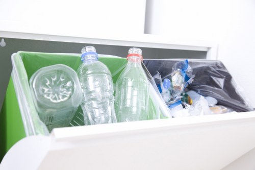 The image displays an open white drawer containing two clear plastic bottles with blue and red caps, both partially visible, resting in a green plastic bin. Next to the bottles, part of a black rubbish bag filled with various discarded items can be seen, including plastic wrappers and crumpled packaging. The background shows a light, clean environment, possibly part of a kitchen or waste disposal area, with the focus on the waste materials ready for collection. The setting emphasizes the typical presentation of domestic or commercial waste awaiting professional rubbish removal services, such as those provided by Commercial Waste Removal Penge. The materials and colors of the bottles, packaging, and bin are clearly visible, with details suggesting they are plastic and the bag's contents are assorted waste materials. The scene illustrates waste items organized in a container, ready for disposal or collection, reflecting routine rubbish management practices in a British property.