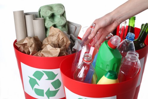 A close-up image of two red recycling bins with a white recycling symbol on each, filled with various household waste materials. The left bin contains several brown crumpled paper bags and a green cardboard egg carton, with some cardboard tubes visible behind them. The right bin holds an assortment of plastic bottles and containers, including a transparent water bottle, a green detergent bottle, and various beverage bottles in different colours like red, green, and clear. A human hand, wearing a clear plastic glove, is seen reaching into the right bin, indicating sorting or collection activity. The background is plain and neutral, focusing attention on the waste items within the bins. The setting suggests a waste collection or recycling process, relevant for a rubbish removal service in the Penge area, where proper waste segregation is emphasized, aligning with the services offered by Commercial Waste Removal Penge.