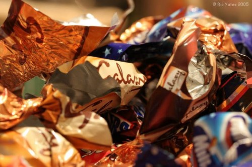 A close-up photograph of a large collection of crumpled metallic and foil wrappers, predominantly in shades of copper, silver, and multicoloured patterns, piled together in a disorganized manner. The wrappers exhibit glossy, reflective surfaces with some displaying partially visible text and branding, indicating that they are from confectionery or snack packaging. The background is out of focus, emphasizing the shiny, irregularly shaped wrappers in the foreground, which are situated on a flat surface such as a table or floor. The image context suggests waste or rubbish, and it relates to rubbish removal services by highlighting the types of waste materials that might be collected and disposed of during a professional rubbish clearance in the Penge area. Overall, the composition emphasizes the textures and colors of discarded packaging materials under natural or artificial lighting, suitable for use on a commercial waste removal website focusing on rubbish collection and disposal services.