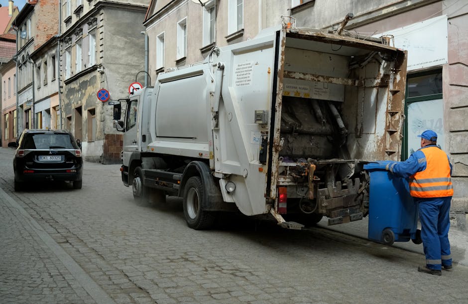 A person is seen disposing of crumpled brown paper bags into a grey waste bin with an open lid, situated on a wooden surface. The person's arm, partially visible, is reaching into the bin to place a p